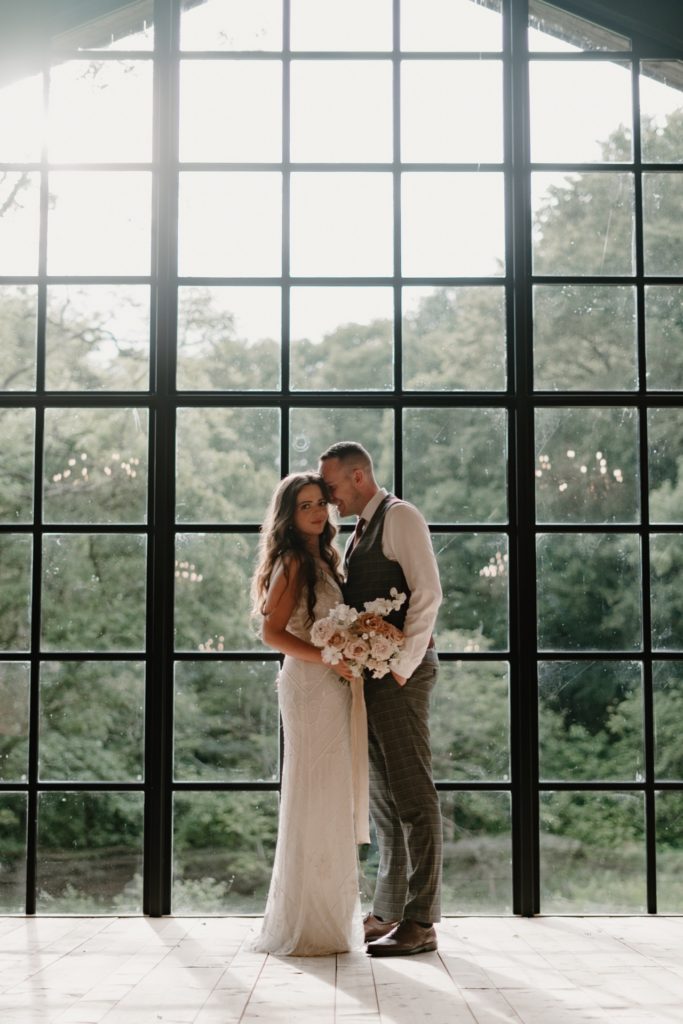 Bride and groom snuggled together standing in front of large window