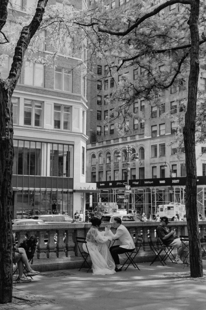 A couple on their wedding day sit at a small round table outside the New York Public Library.
