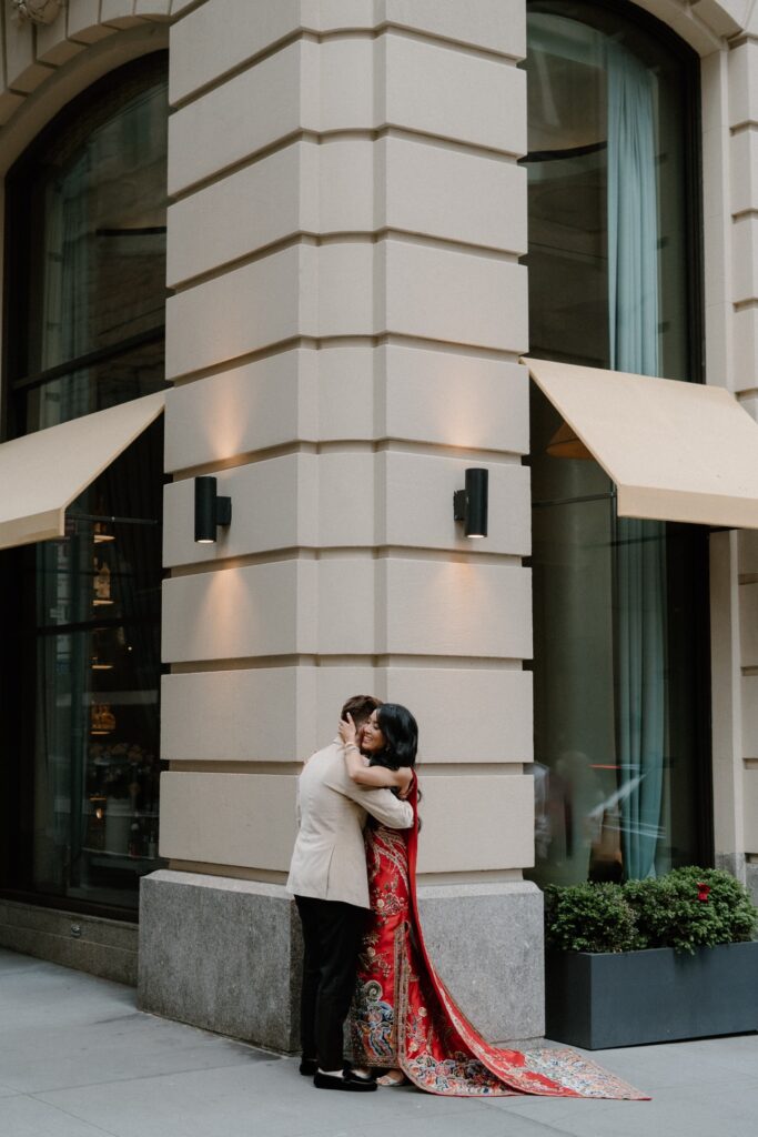 A couple on their wedding day embrace after their first look outside The Wall Street Hotel. One bride wears a velvet tux jacket and the other wears a traditional red Chinese dress