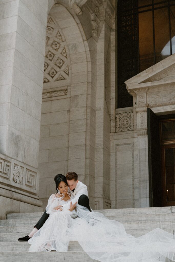 Couple sit on the steps of the New York Public Library. One bride wears a white tux and the other a Galia Lava gown and veil which flows down the steps.