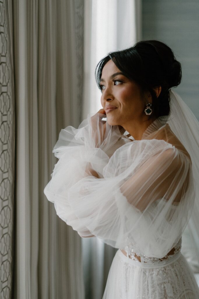 Bride wearing Galia Lava wedding dress stands in front of a window putting in her earring.