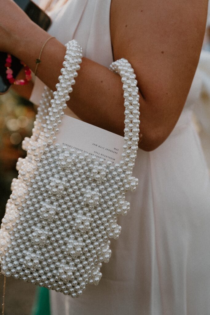 A pearl handbag with an order of service poking out of the top on the arm of a woman in a satin dress
