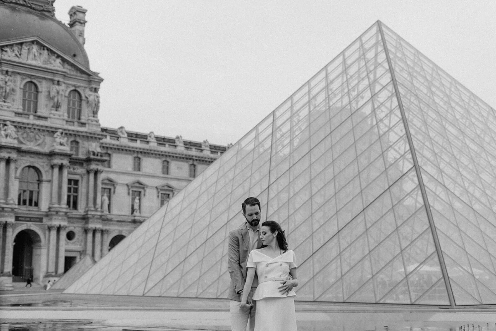 Couple stood cuddled together in front of one of the glass pyramid of the Louvre.