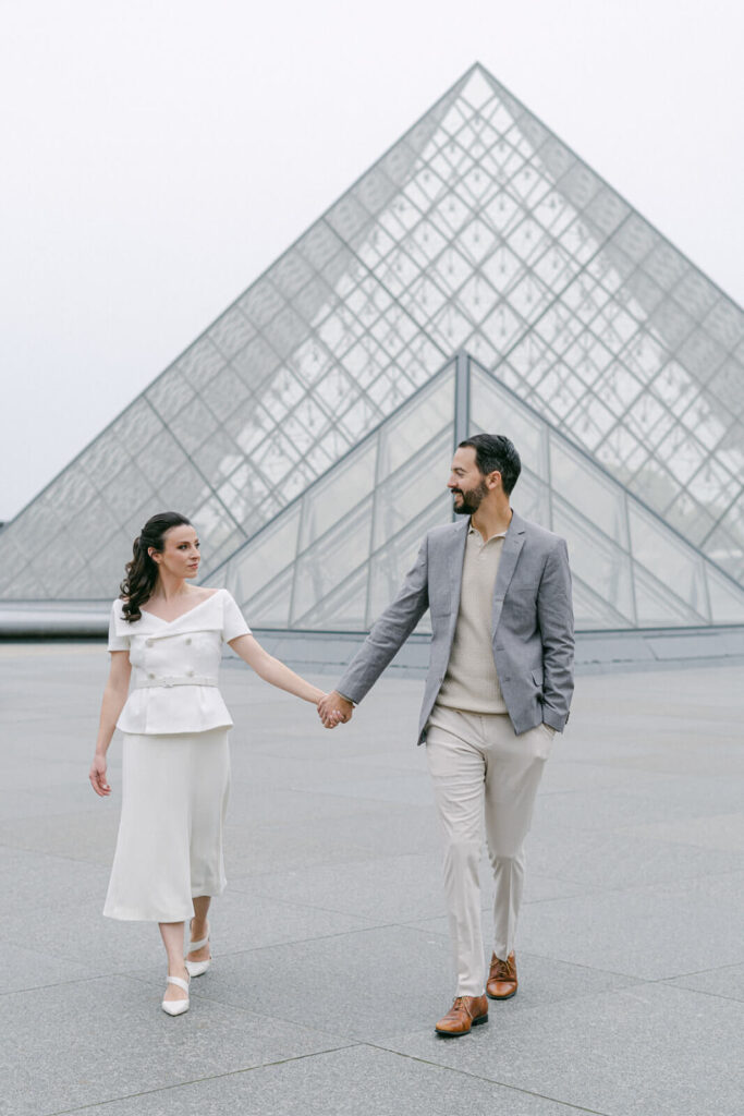 Couple walking hand in hand in front of the Louvre in Paris.