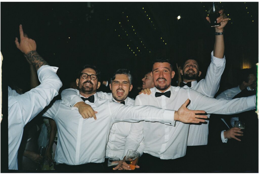 A group of men in black tie with their arms around each other, looking directly at the camera and singing on the dance floor at a wedding